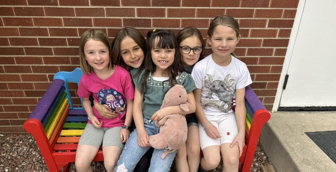 A group of elementary school aged children sit on a rainbow-painted bench in the school playground area.